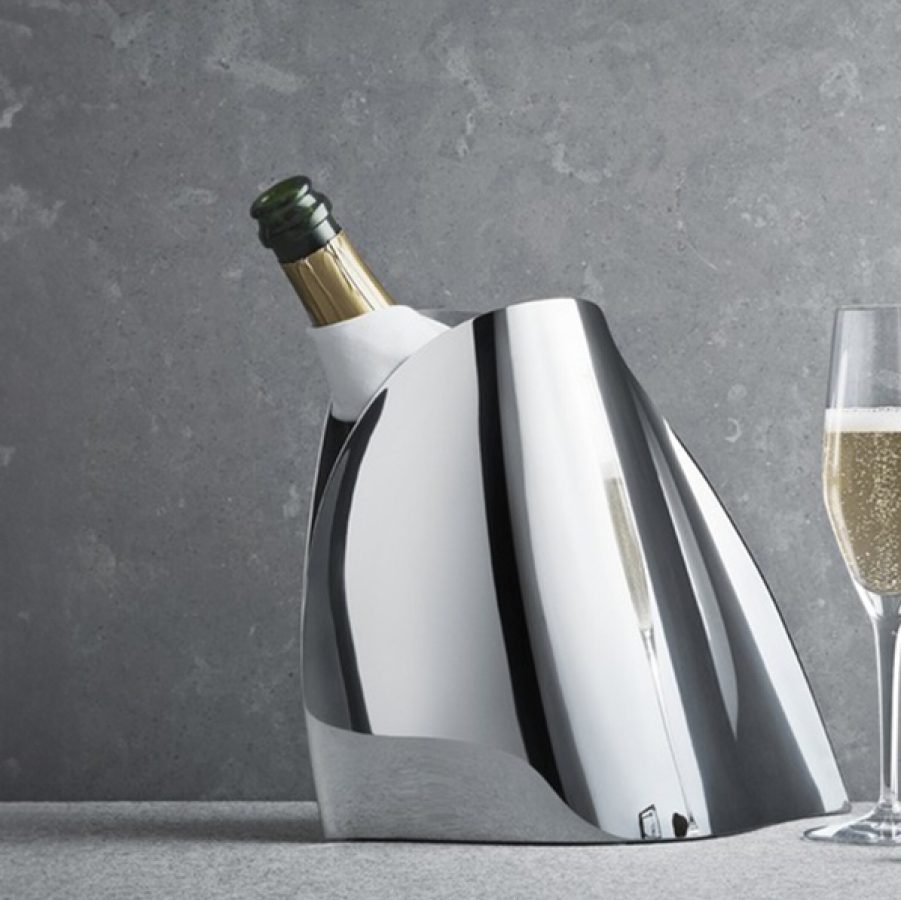 An opened bottle of champagne in a Georg Jensen stainless steel champagne cooler bucket next to a glass of champagne