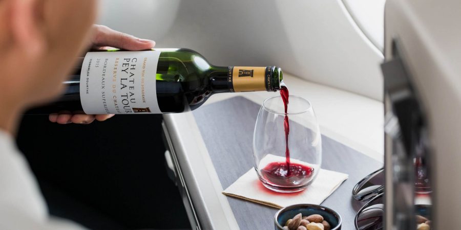 Een hand schenkt rode wijn in een wijnglas zonder steel op een plateau bij het raam van een stoel in Cathay Pacific Business Class. Ernaast staat een kleine kom amandelen.