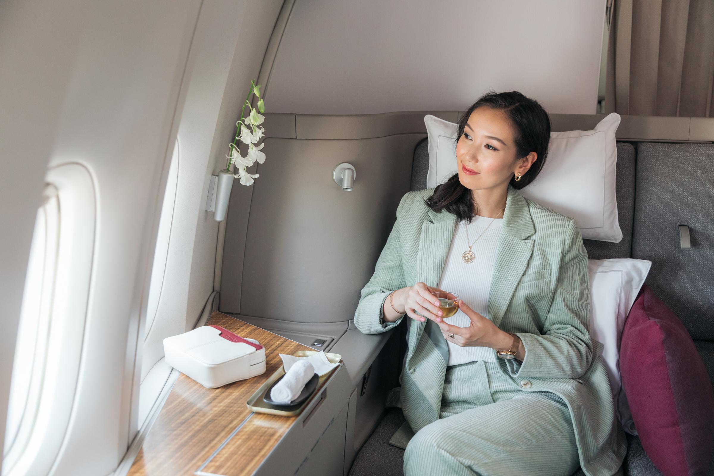 An Asian passenger in a Cathay Pacific First class seat, with two white pillows cushioned behind her. Smiling, she is holding a cup of tea.