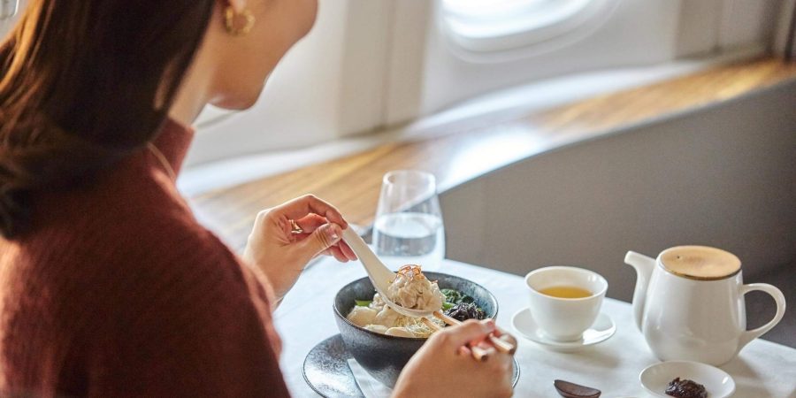 在國泰航空的頭等客艙內，一名女士正在用筷子和湯匙吃一碗餛飩面，旁邊放著中式調味料、一杯茶和一杯水。 