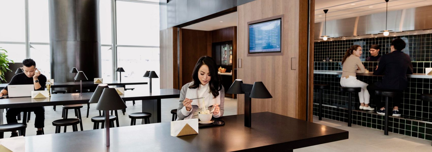  A female passenger is sitting at a long table with chopsticks in her left hand eating a bowl of noodles inside a Cathay Pacific lounge.