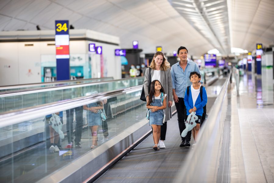 A family of four with two children on a moving walkway in the Hong Kong International Airport. The boarding gate signs can be seen in the background.