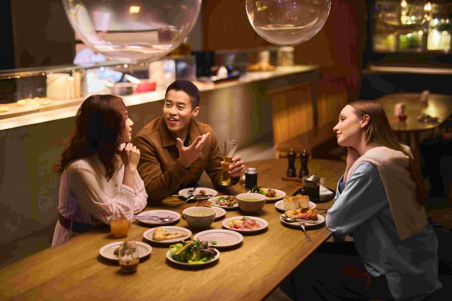 Three diners sitting down at a table with a full spread of food in front of them. They are engaged in conversation.