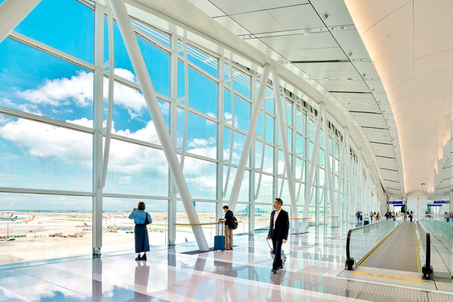 The Sky Bridge walkway from inside Hong Kong International Airport. The floor-ceiling-windows look out to the tarmac and blue skies above.